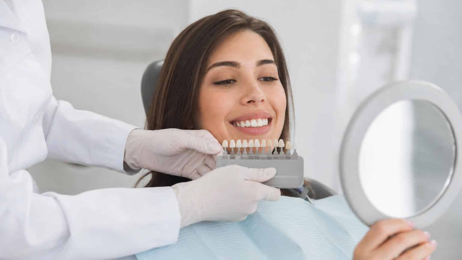Dentist holding a tooth shade guide against a smiling woman’s teeth to select the perfect color for ceramic veneers. Dentist holding a tooth shade guide against a smiling woman's teeth to select the perfect color for ceramic veneers.