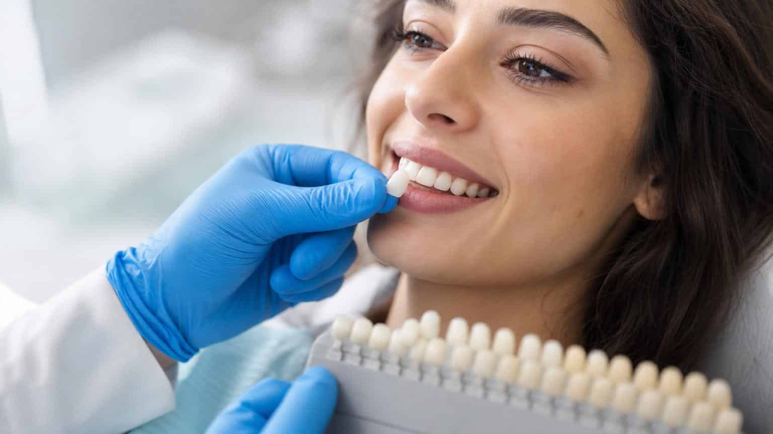 Dentist in blue gloves matching a resin veneer shade to a smiling woman’s natural teeth using a dental color guide. Dentist in blue gloves matching a resin veneer shade to a smiling woman's natural teeth using a dental color guide.