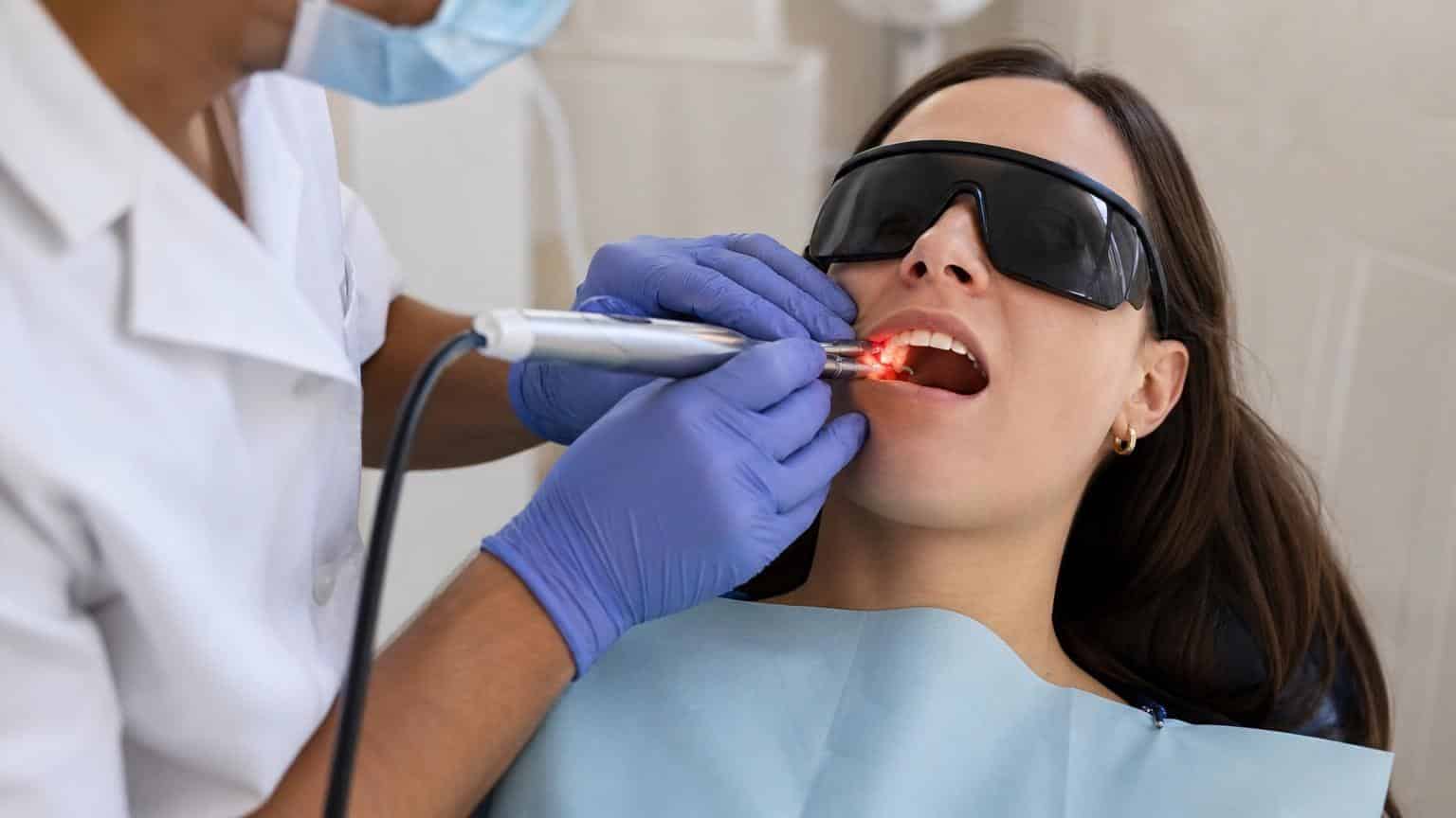 Dentist using a handheld laser tool with a red light to treat the gums of a patient wearing protective black goggles. Dentist using a handheld laser tool with a red light to treat the gums of a patient wearing protective black goggles.